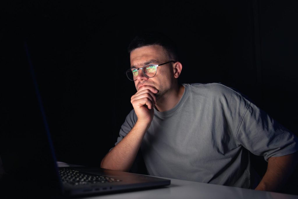 Hombre con gafas mirando pensativo la pantalla de un portátil en una habitación a oscuras